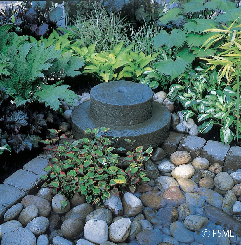 Mill stone among pebbles and surrounded by tranquil plantings of ajuga, heuchera, gunnera and hosta.